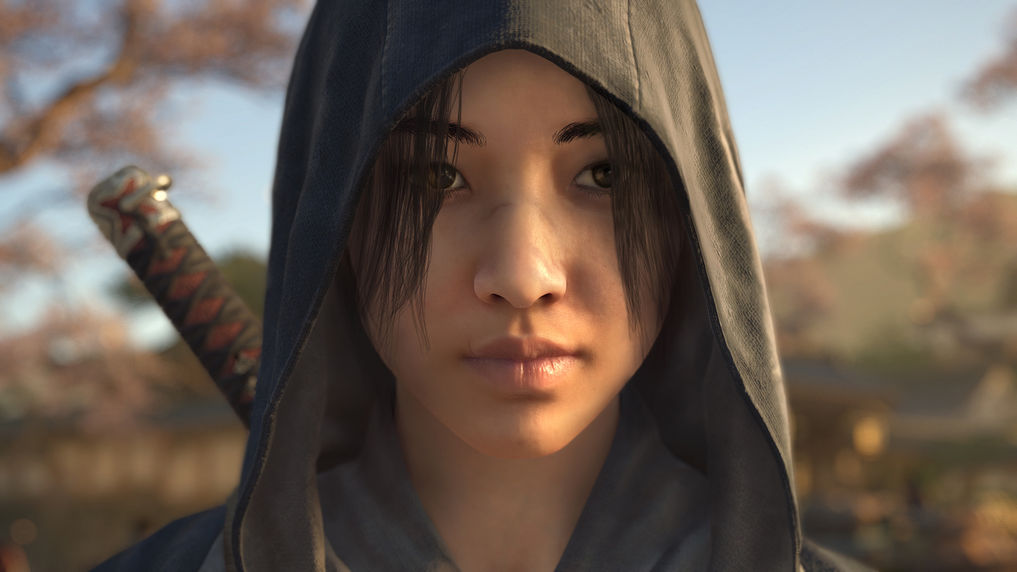 In this screenshot from Assassin’s Creed Shadows, the character Naoe is seen in close-up. She is wearing a hood, carrying a sword on her back, and standing outdoors. 