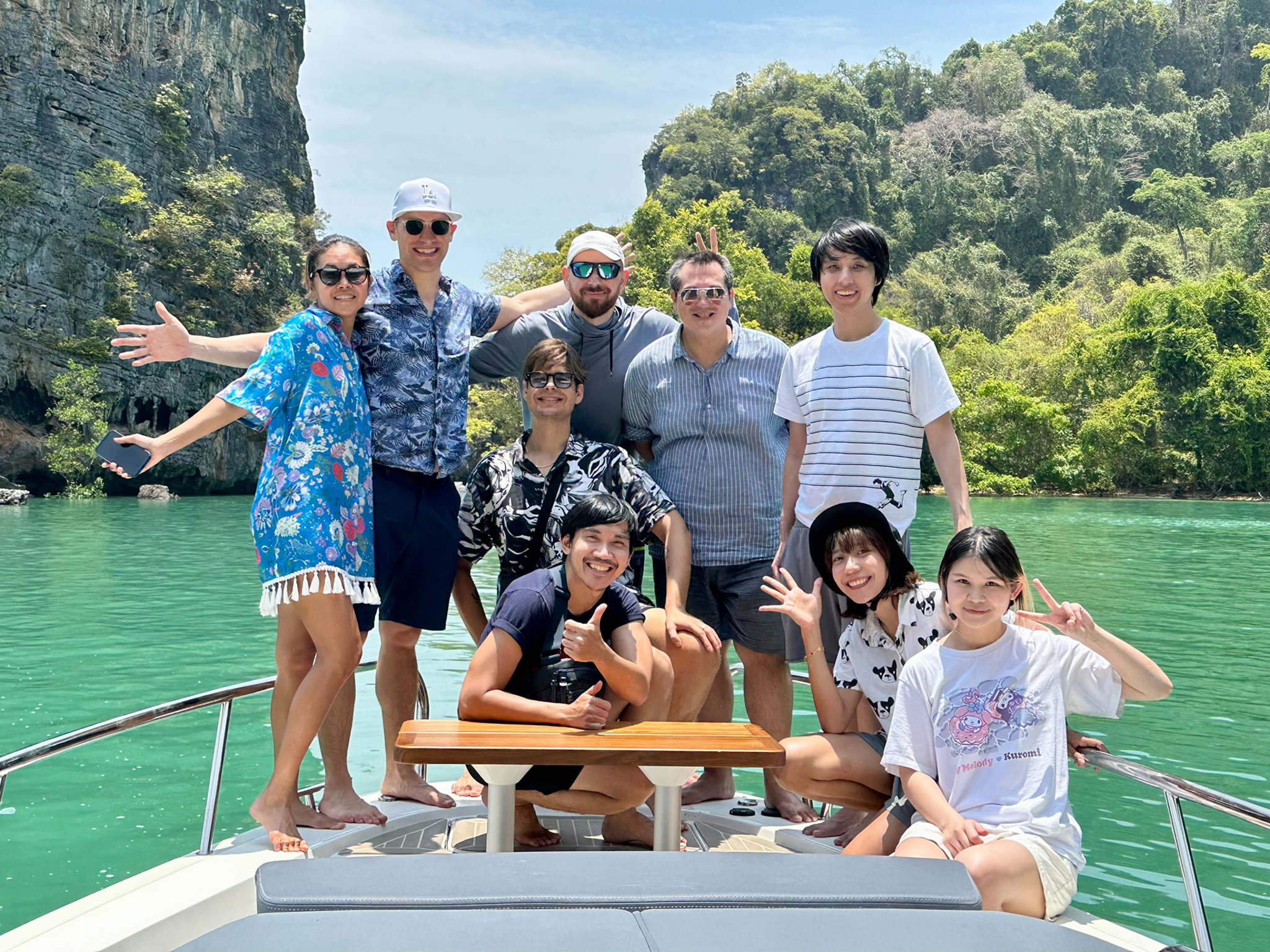 A group photo of nine people posing cheerfully on the bow of a ship. They are dressed in casual warm-weather clothing — shorts, T-shirts, and a floral wrap. Several people are waving, making peace signs, or giving thumbs-up. Behind them, dramatic green limestone cliffs rise above vivid turquoise water under a bright blue sky.