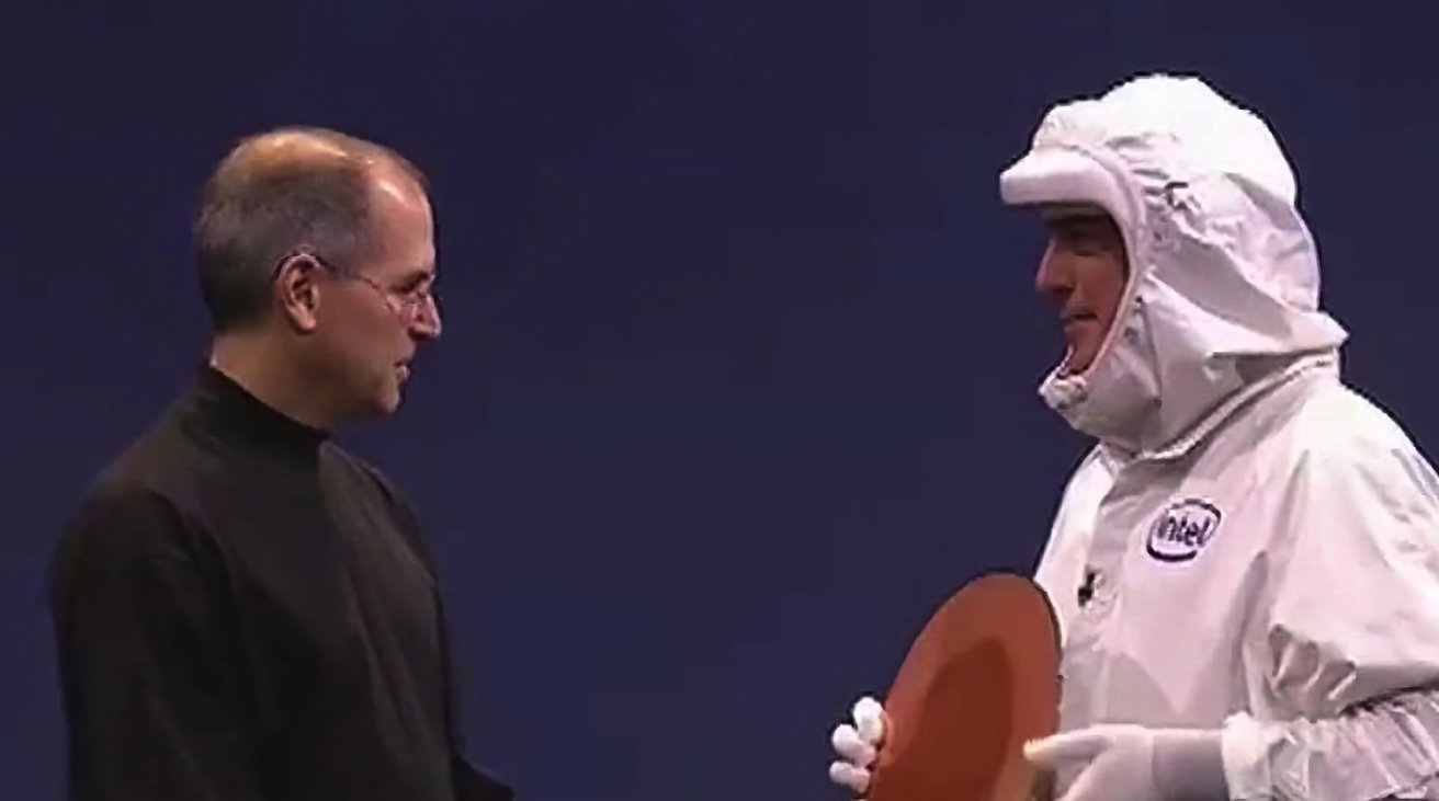 Two men on stage: one in a black turtleneck, the other in a white Intel cleanroom suit holding a large circular silicon wafer against a plain dark background