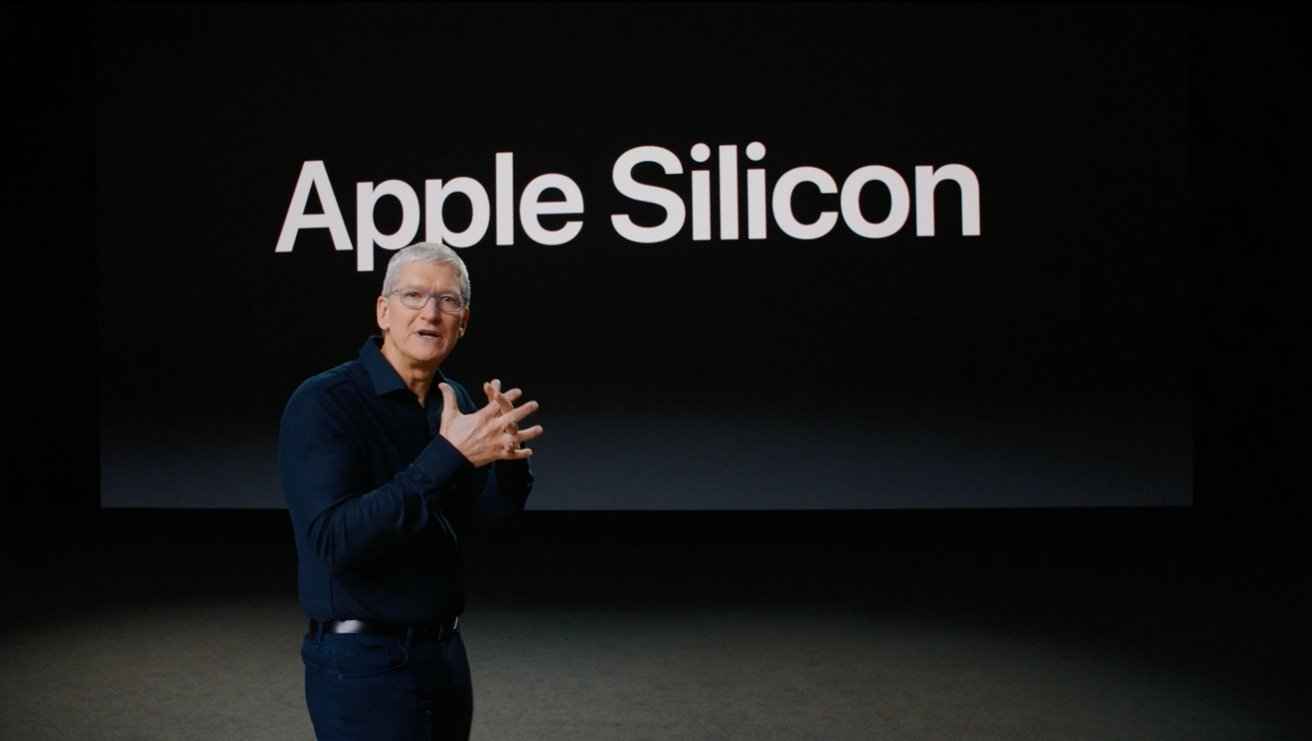Man in dark clothing speaking onstage in front of a large black screen displaying the white text Apple Silicon