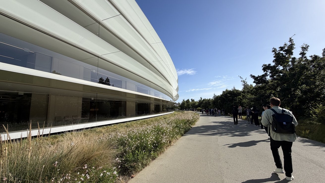 Curved modern glass building beside a landscaped path with grasses and flowers, people walking in sunlight under a clear blue sky, some carrying backpacks near trees