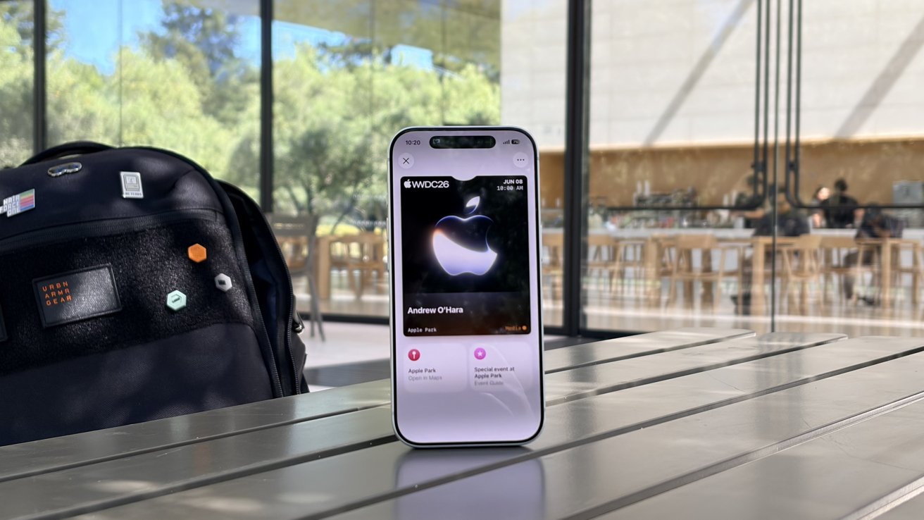 Smartphone standing on an outdoor cafe table, displaying a dark Apple-themed music screen, with a backpack on the left and modern glass-walled building and trees in the background