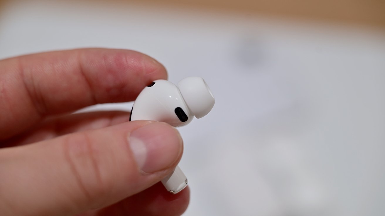 Hand holding a single white wireless earbud with silicone tip, shown close up against a softly blurred light background