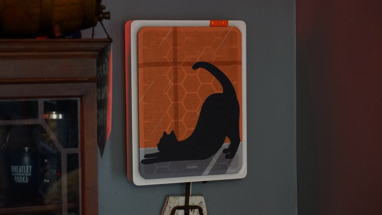 Artwork on an air purifier showing a black cat stretching against an orange geometric background, with part of a wooden cabinet visible to the left and an electrical outlet below