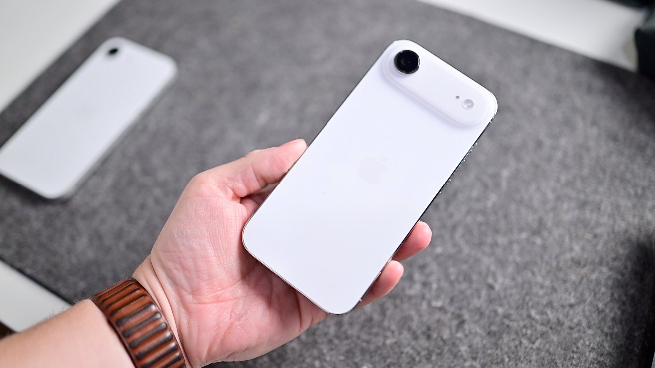 Hand holding a white smartphone with a single rear camera, viewed from the back, over a gray fabric surface, with another similar phone lying flat in the background